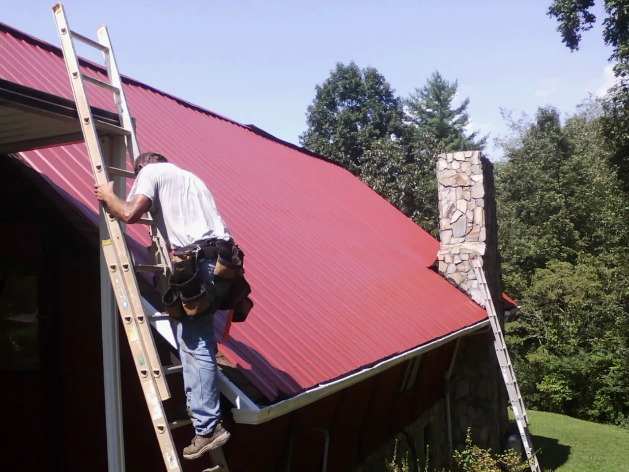 Roofer on a ladder beside a red metal roof and stone chimney — Soffit & Fascia Repair by Roofing Brothers in Suffolk